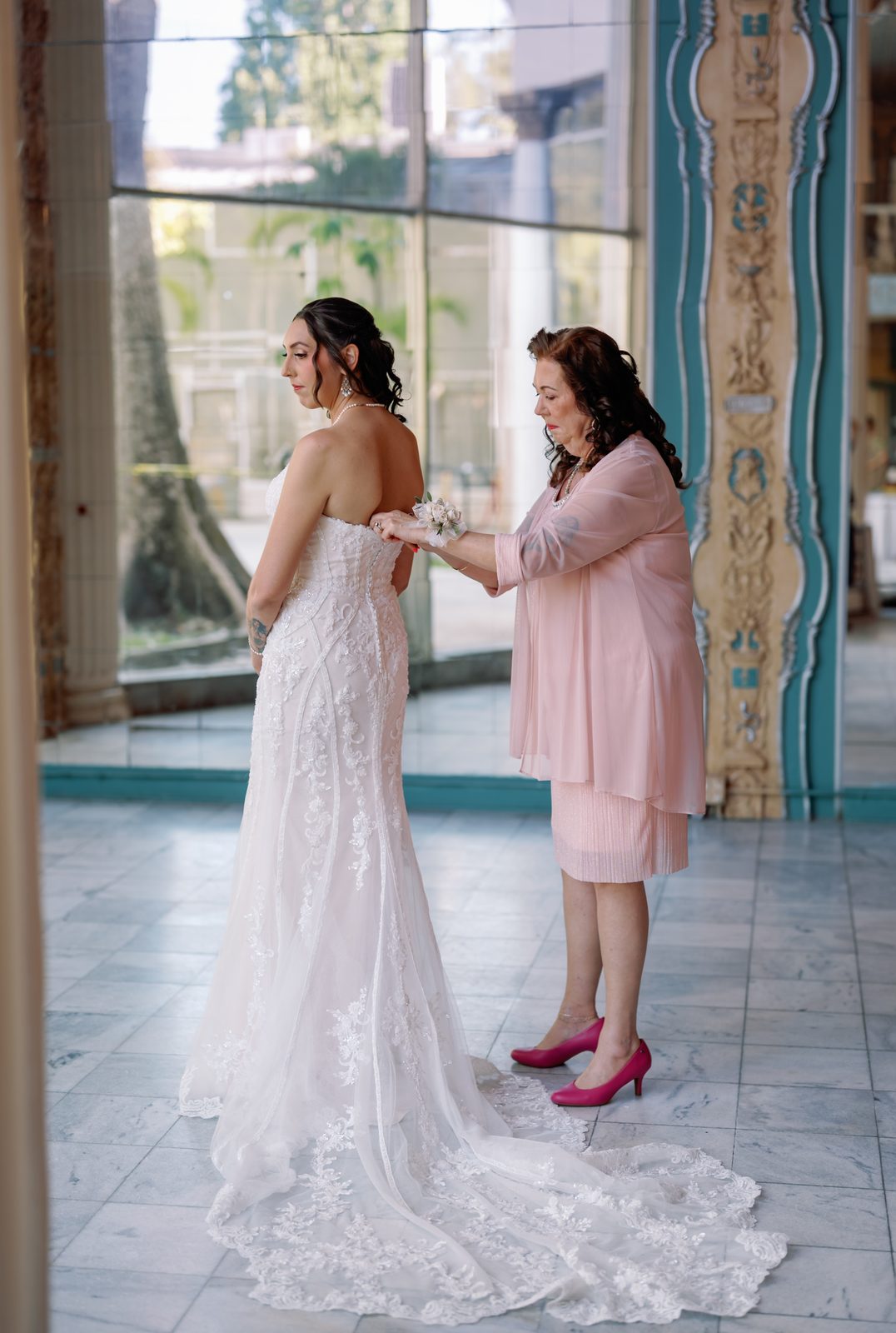 A bride's mother fastening the back of her dress in a sunlit room before the ceremony