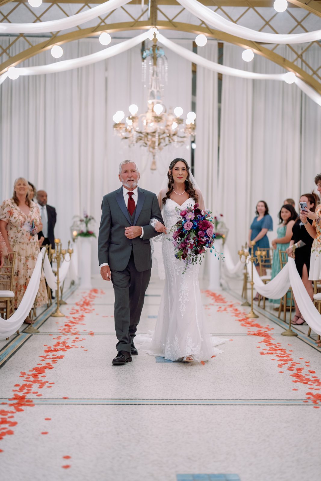 A father walking his daughter down a petal-strewn aisle toward the ceremony